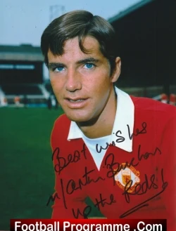 Footballer in a red kit signs a football programme on a stadium pitch, with handwriting across his chest.