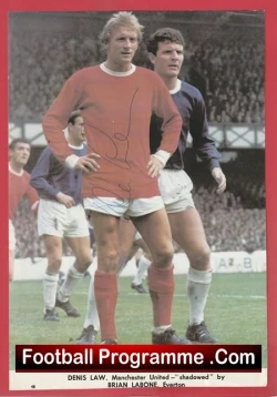 Two soccer players in the foreground during a match, one in a red shirt and white shorts and the other in a dark kit, with a 'Football Programme .Com' banner at the bottom.