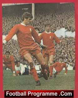 Action shot of football players in red kits on a green pitch, crowd in the stands, with a 'Football Programme.Com' banner at the bottom.